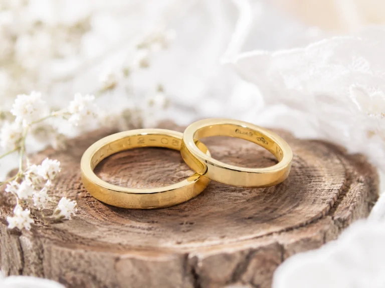 Two classic yellow gold wedding rings resting on a wooden log, surrounded by white flowers in a natural wedding setting.