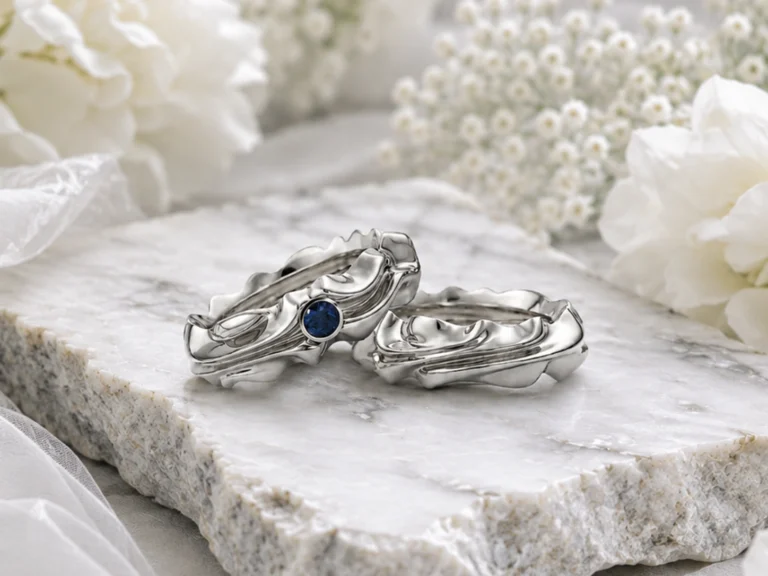 A pair of narrative wedding rings in white gold with a blue sapphire, photographed on natural stone amongst white flowers.